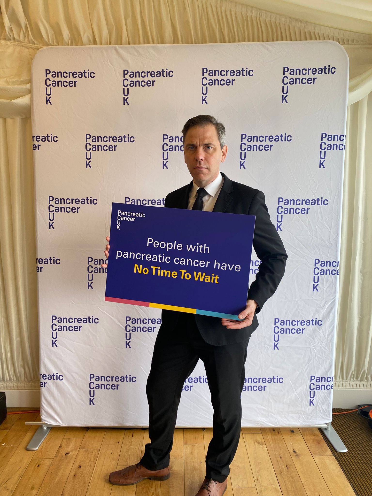 Chris Evans MP is holding a sing that reads 'people with pancreatic cancer have no time to wait'