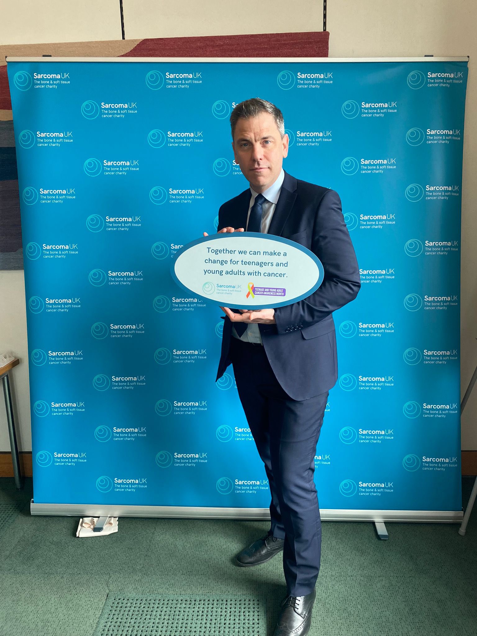 Chris Evans MP is holding a light blue sign with dark blue lettering, that reads: together we can make a change for teenagers and young adults with cancer